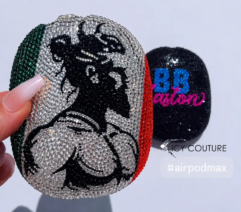 A close-up of a hand holding a rhinestone-embellished oval airpod case featuring a black silhouette graphic over green, white, and red vertical stripes. A second black airpod case with blue and pink lettering sits behind it on a plain light background.